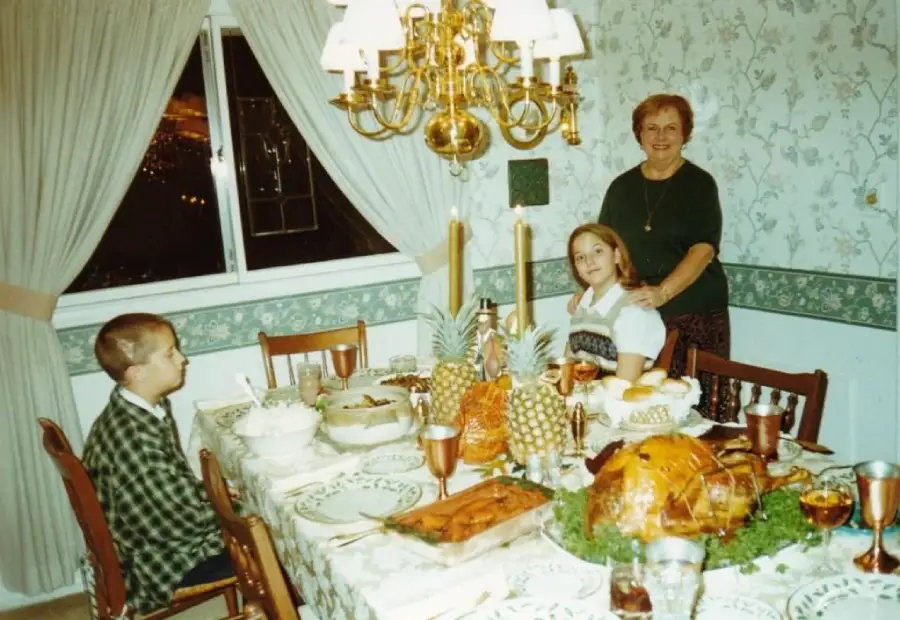A boy and a girl sit at a dining table with various dishes and a large turkey, while an older woman stands behind them, all smiling in a warmly lit room with patterned wallpaper.