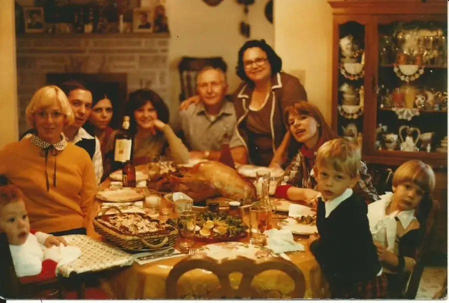 A family gathers around a dining table set with food, including a roast turkey. Adults and children pose and smile in a warmly lit room with a china cabinet in the background.