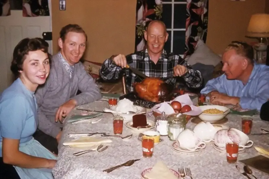 Four adults sit around a table set for a meal, with one person carving a roasted turkey. The table has glasses of juice and various dishes, suggesting a festive or holiday meal in a cozy dining room.