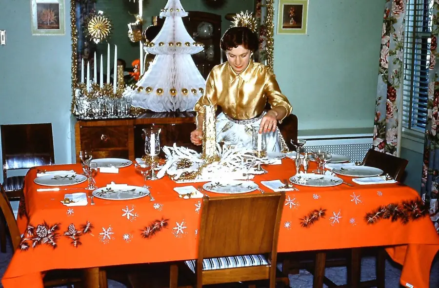 A woman sets a festive dining table with red tablecloth, pinecone and snowflake decorations. A white Christmas tree and candles are on a sideboard in the background.