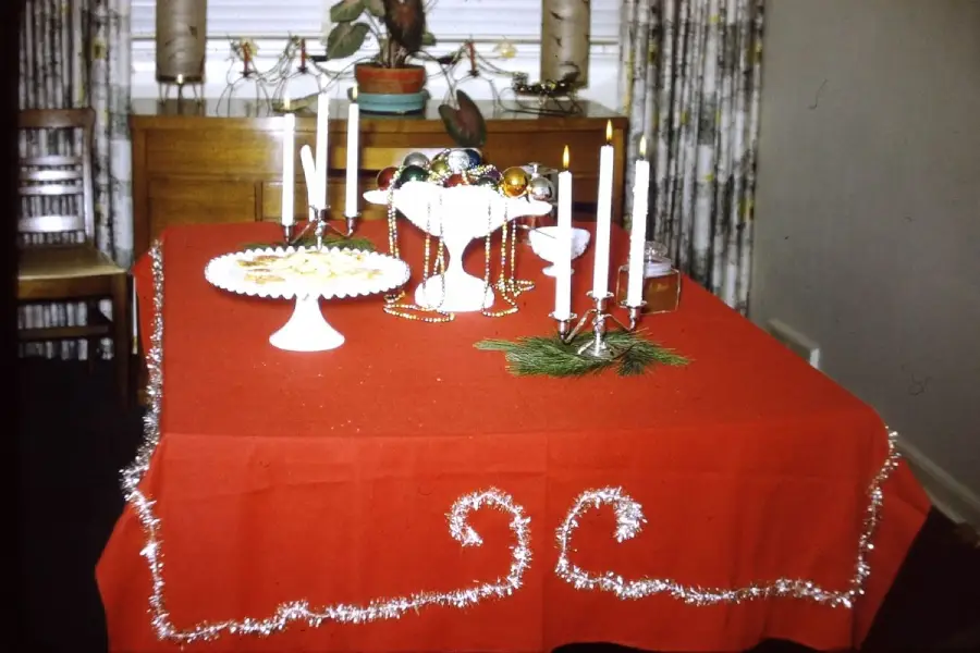 A red tablecloth with silver tinsel decorates a table set with white dishes, candles, pine, and ornaments. The background includes a cabinet, plant, and patterned curtains.