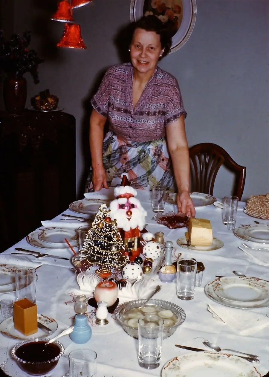 A woman stands smiling behind a decorated dining table with holiday-themed ornaments, glassware, plates, desserts, and cheese, preparing for a festive meal.