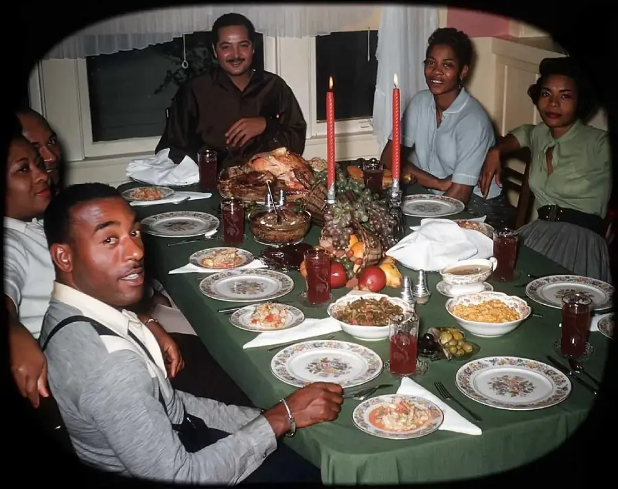 Six people sit around a dining table set with a holiday meal, including turkey, side dishes, fruit, drinks, and two lit candles. Everyone is smiling and dressed neatly, creating a warm, festive atmosphere.