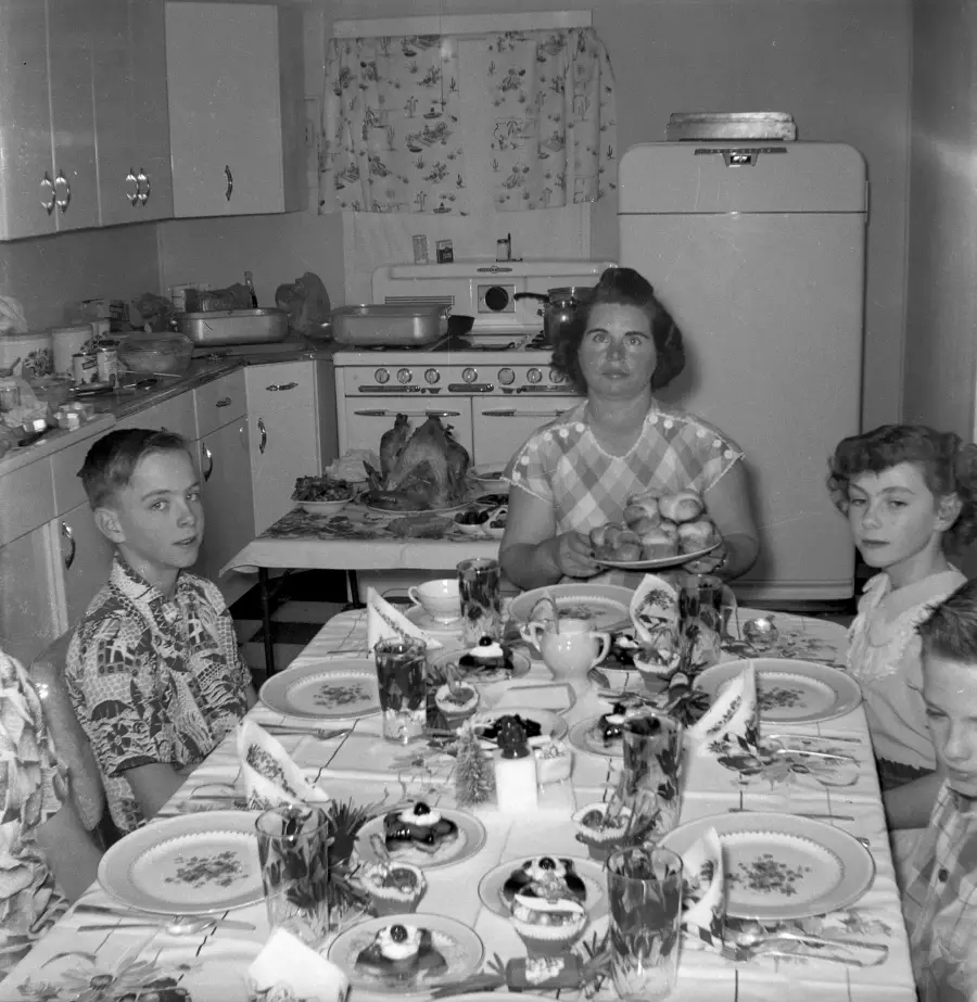 A family of five sits at a kitchen table set with food and desserts. The mother holds a plate of rolls. A turkey is on a tray behind her. The kitchen has floral curtains and 1950s-style appliances.