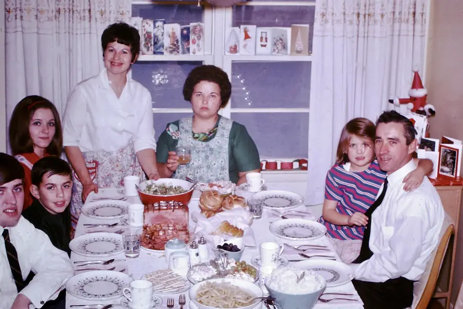 A family of seven poses around a dining table set with food. Vintage decor, Christmas cards, and a Santa decoration can be seen in the background. Most people are smiling; one woman looks serious.