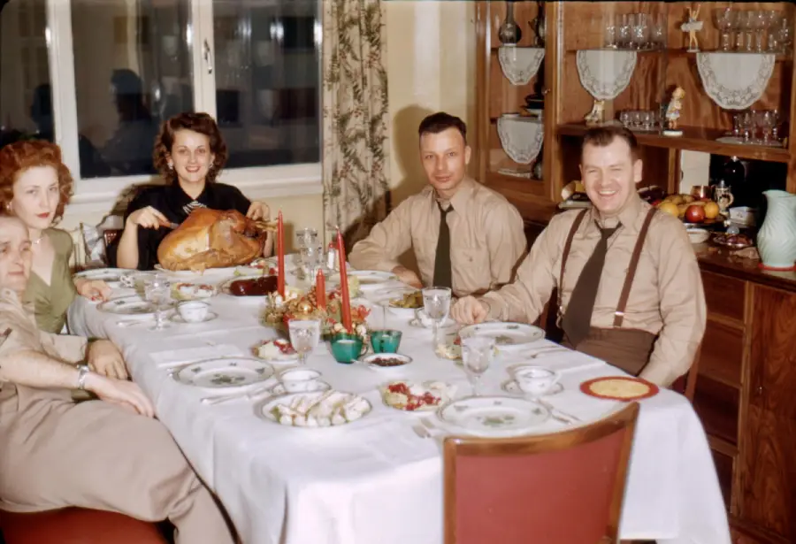 Four adults in vintage attire sit at a dining table set for a festive meal. One woman holds a roast turkey. The table is decorated with candles and dishes. A cabinet with glassware is in the background.