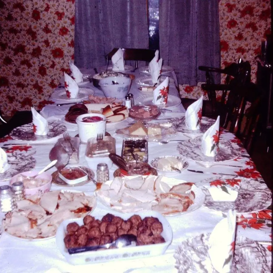 A dining table set for a meal, covered with a white tablecloth, plates, napkins, and various dishes including meatballs, sandwiches, cheese, and desserts, in a room with floral wallpaper and curtains.