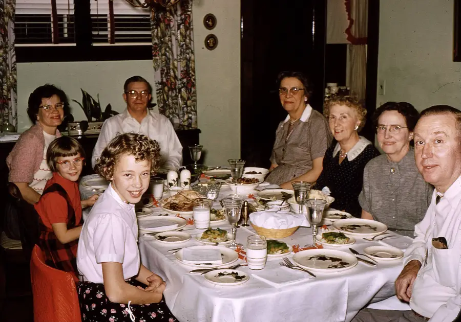 Seven people, including two children, sit around a dining table set with plates and glasses, smiling at the camera in a cozy, warmly lit room with floral curtains and decorations on the walls.