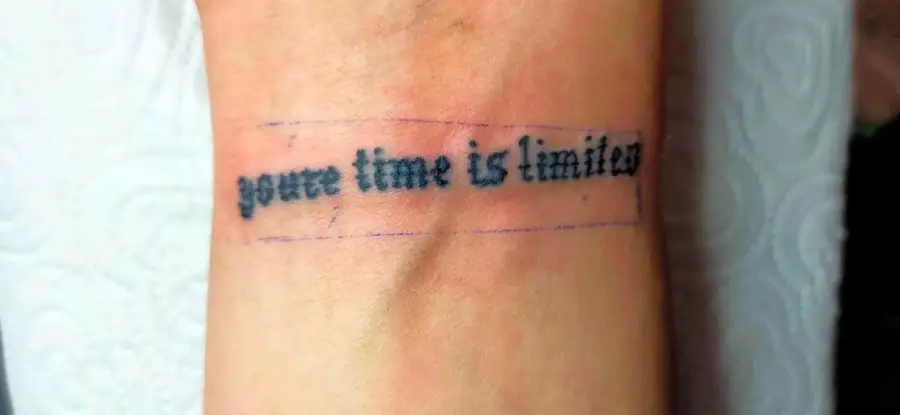 A wrist tattoo with the phrase “your time is limited” in black, typewriter-style font, with a faint rectangular outline around the text.