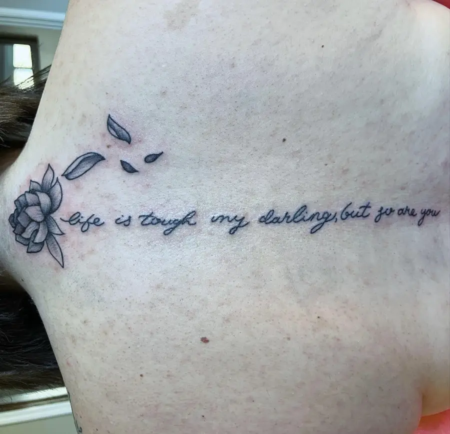 Tattoo of a flower losing petals, with cursive text reading, “life is tough my darling, but so are you,” on the back of a person’s neck.