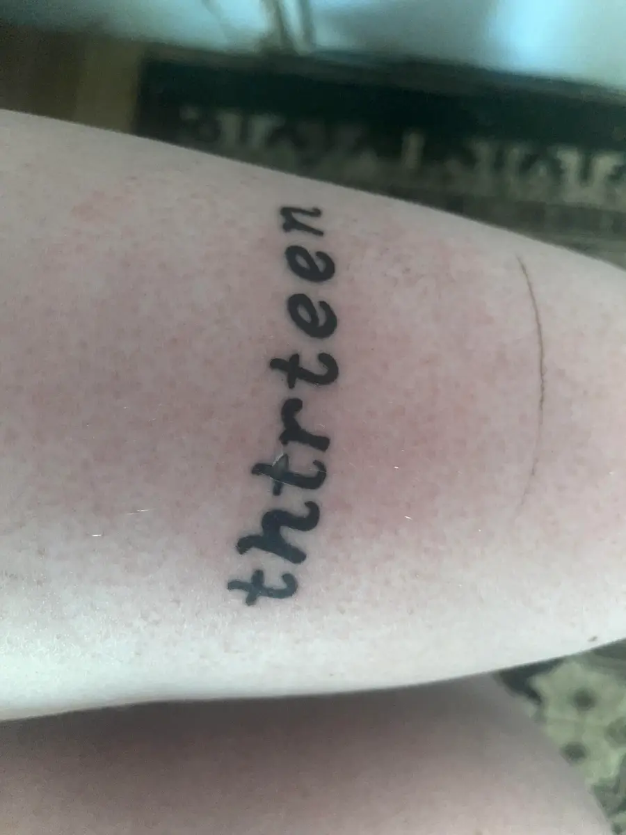 A tattoo on pale skin reads “thirteen” in lowercase cursive letters, but is misspelled as “thrirteen.” The tattoo is freshly done, and the skin appears slightly red around the ink.