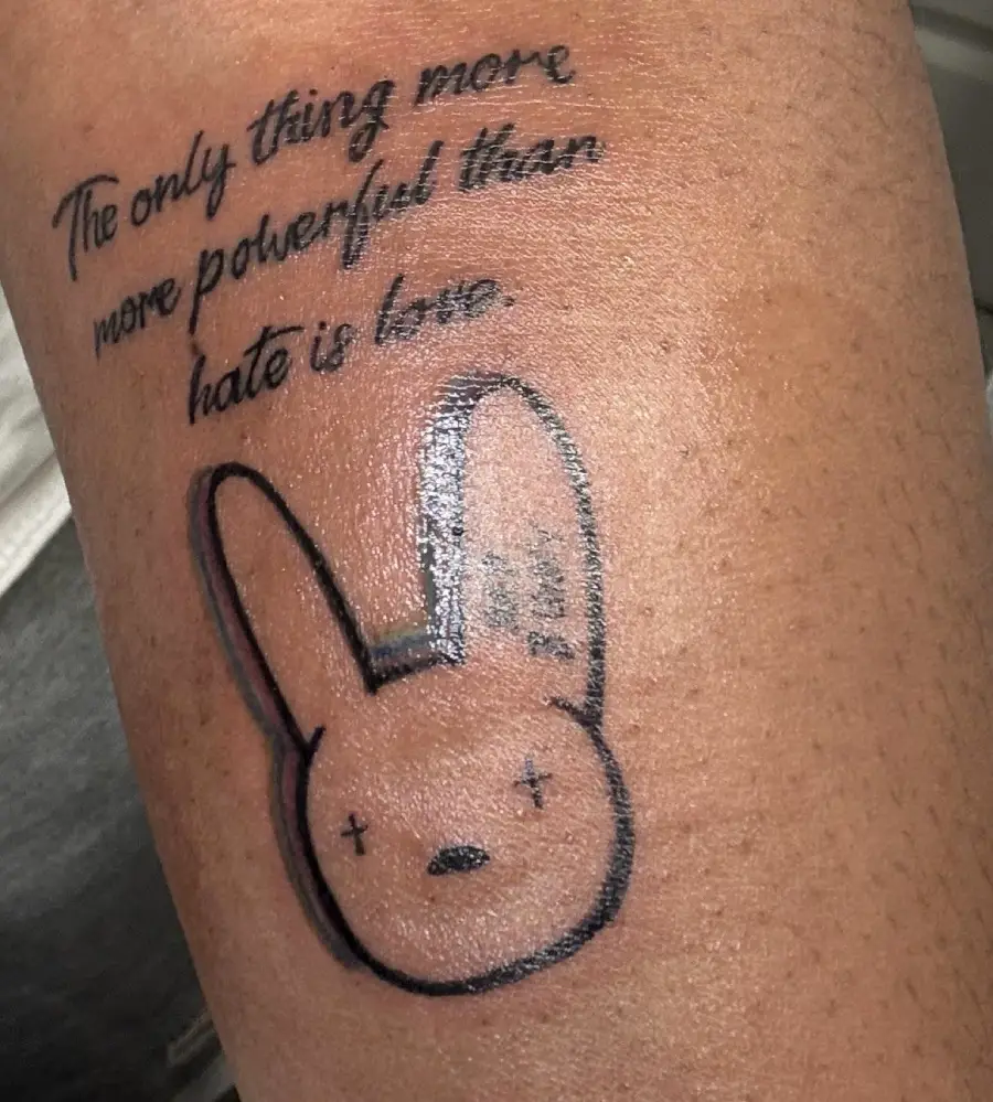 Tattoo of handwritten text above a simple black-outline bunny face with Xs for eyes. The text reads: “The only thing more powerful than hate is love.” Tattoo is on brown skin.