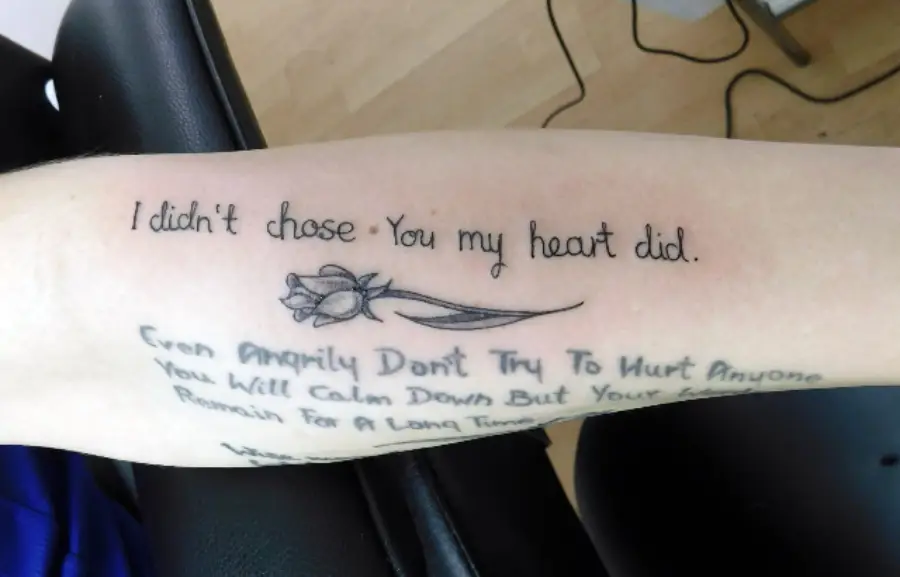 Forearm tattoo reads “I didn’t chose you, my heart did.” with a simple flower design. Another faded tattoo with cursive text is visible below.