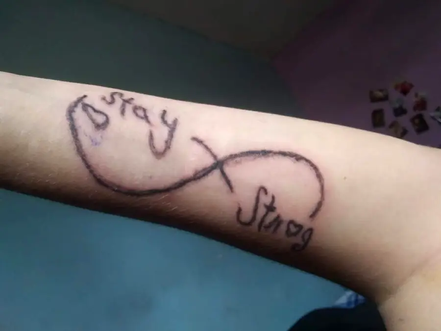 A tattoo on a forearm shows an infinity symbol with the words “stay” and “strong” written in a handwritten style.