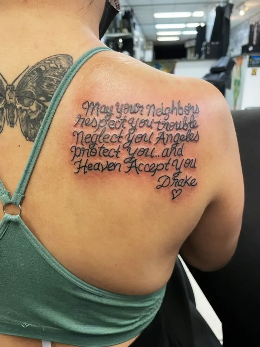 A person with a butterfly tattoo on their shoulder blade has cursive script tattooed below it, featuring a quote and a small heart at the end. The individual is wearing a green top with thin straps.