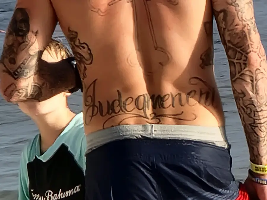 A person with multiple tattoos, including large script text “Judgement” across their lower back, stands near a child by the water. The child is wearing a two-tone shirt and a cap.