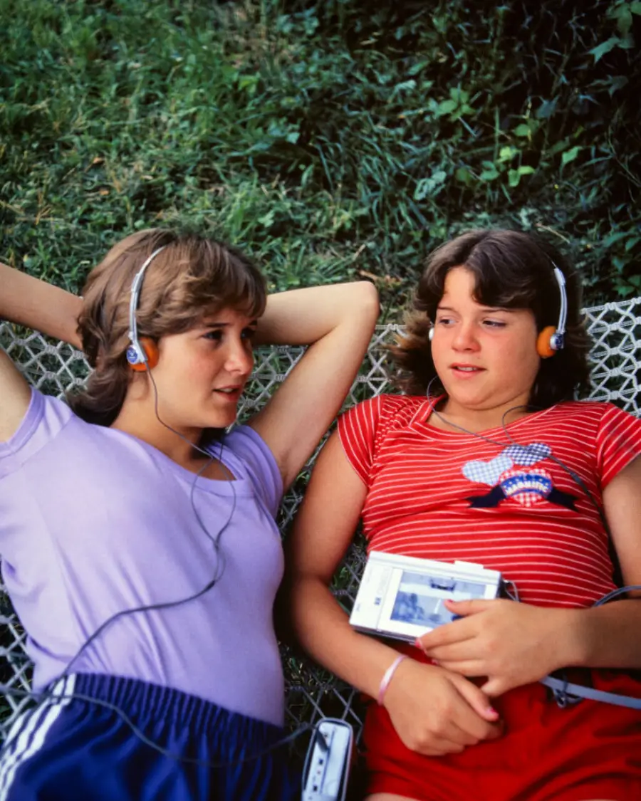 Two teenagers lie on a net outdoors, wearing headphones and casual clothes, listening to a portable cassette player together. Grass is visible in the background.