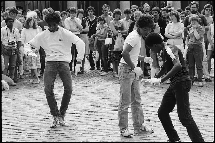 Three young men breakdancing on a cobblestone street, wearing casual clothes and gloves, while a crowd of people watches them perform.