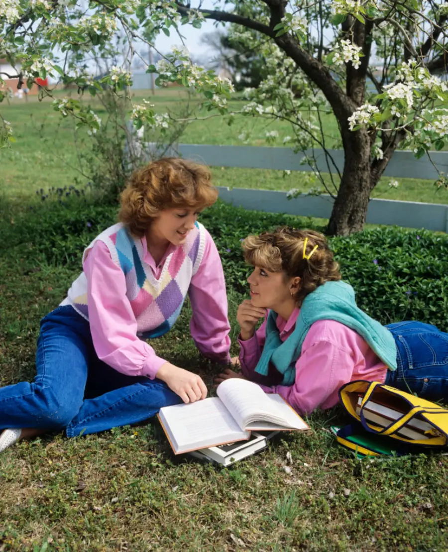 Two young women sit on grass under a flowering tree, studying an open book together. Both wear jeans and pink shirts; one has a green sweater tied around her shoulders. A backpack and notebook lie nearby. It is a bright, sunny day.