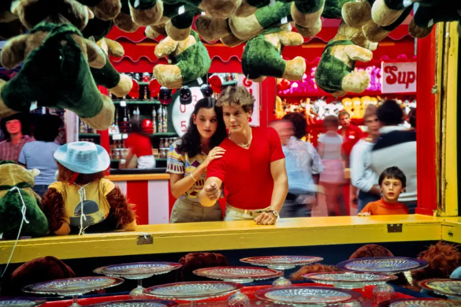 A couple stands close together at a carnival game booth with hanging plush frogs, concentrating on the game. People and colorful lights blur in the busy background. A young boy watches from the side.