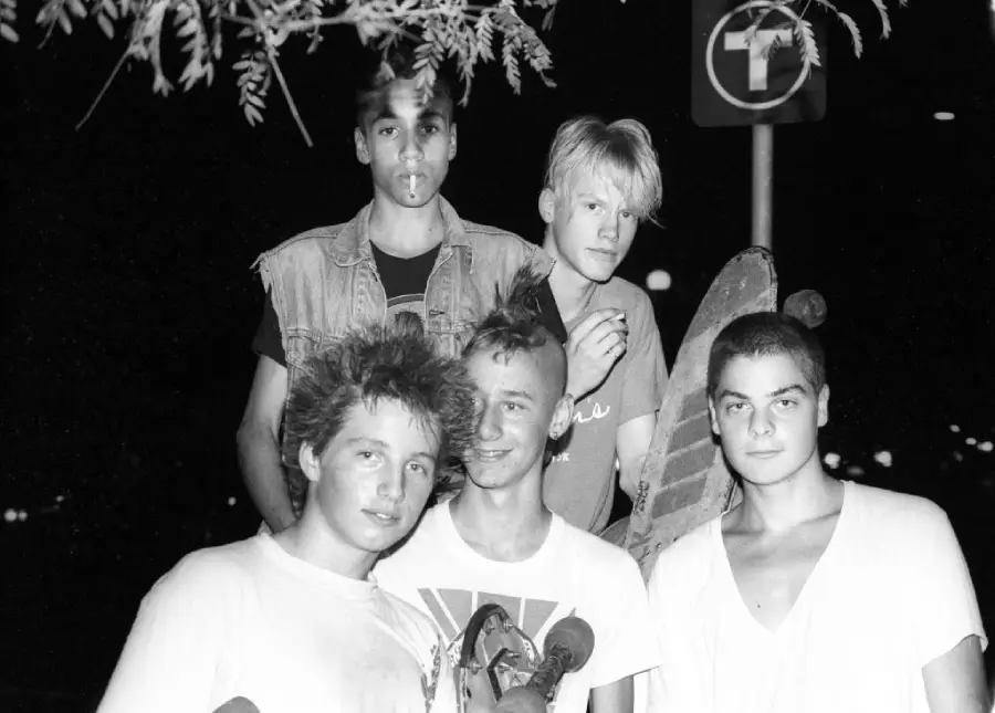 Five teens with skateboards pose at night under a tree branch. One wears a denim vest, one has a mohawk, and a street sign with a T symbol is visible in the background.