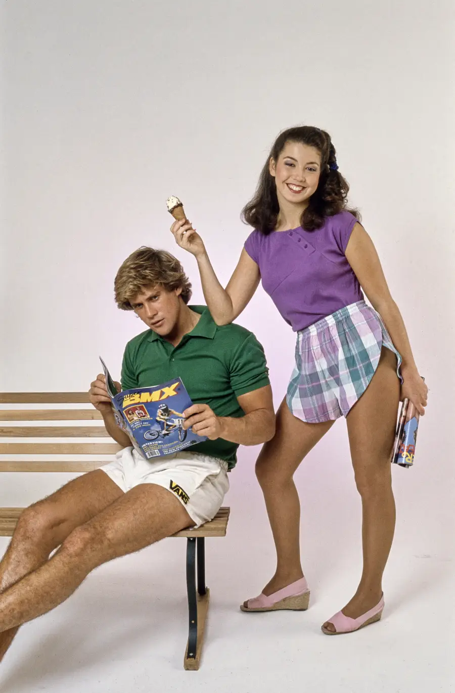 A man in a green shirt and white shorts sits on a bench reading a comic, while a woman in a purple top and plaid skirt stands smiling, holding an ice cream cone and a rolled-up magazine.
