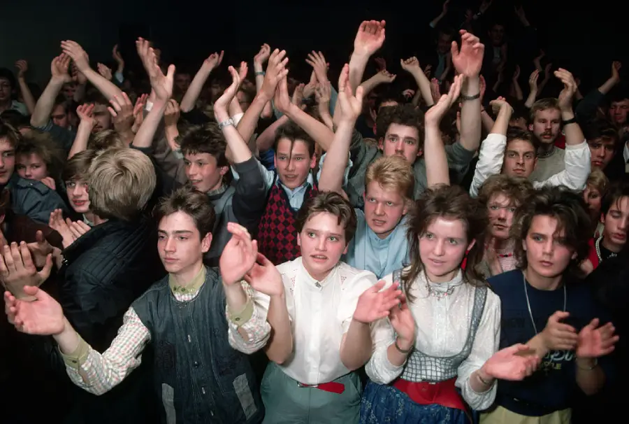 A crowd of young people enthusiastically clapping with raised hands at a concert or event, wearing 1980s-style clothing, with excited and cheerful expressions.