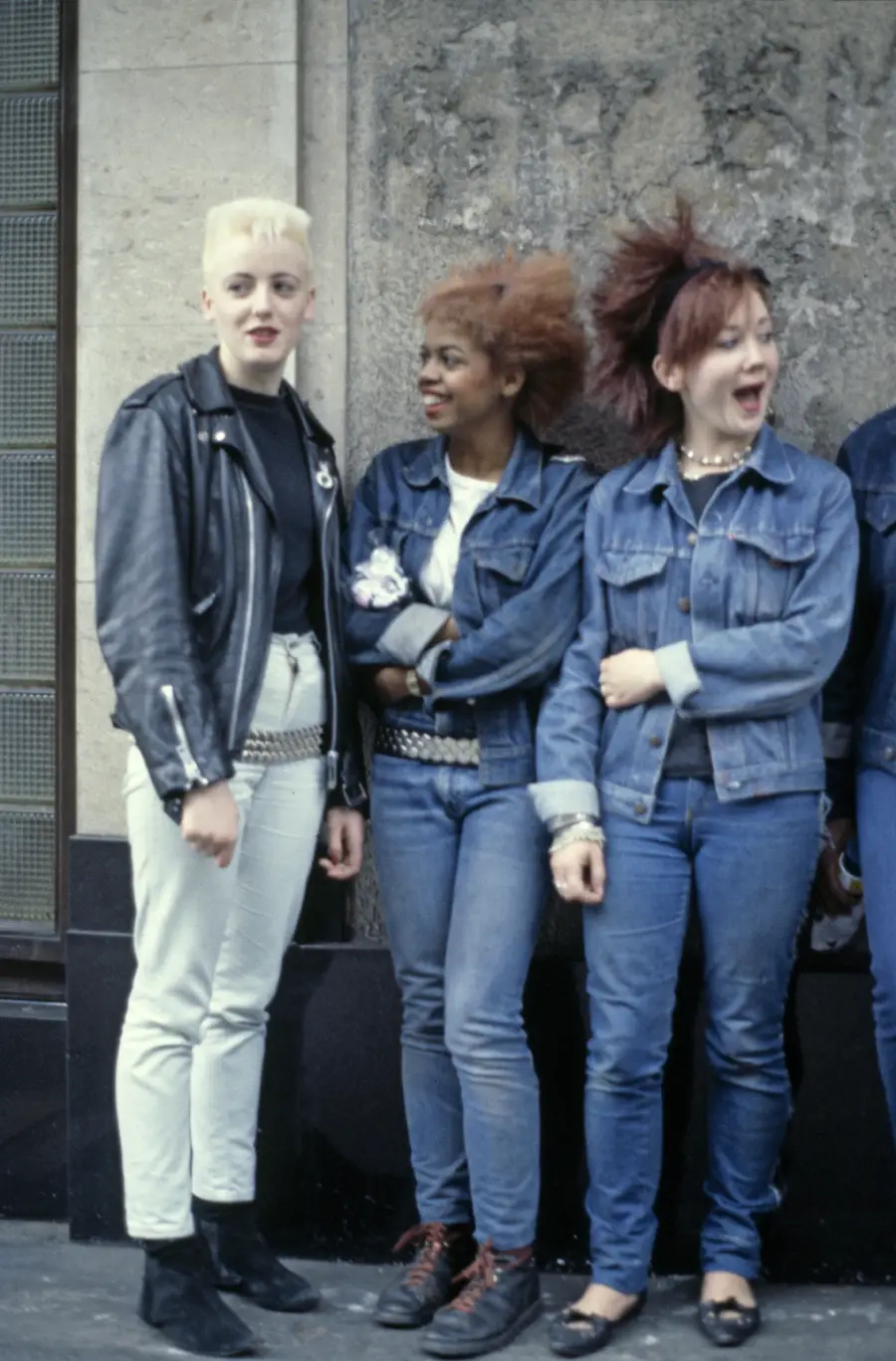 Three young women with punk hairstyles and outfits stand against a stone wall, wearing denim and leather jackets. They appear to be chatting and laughing together.