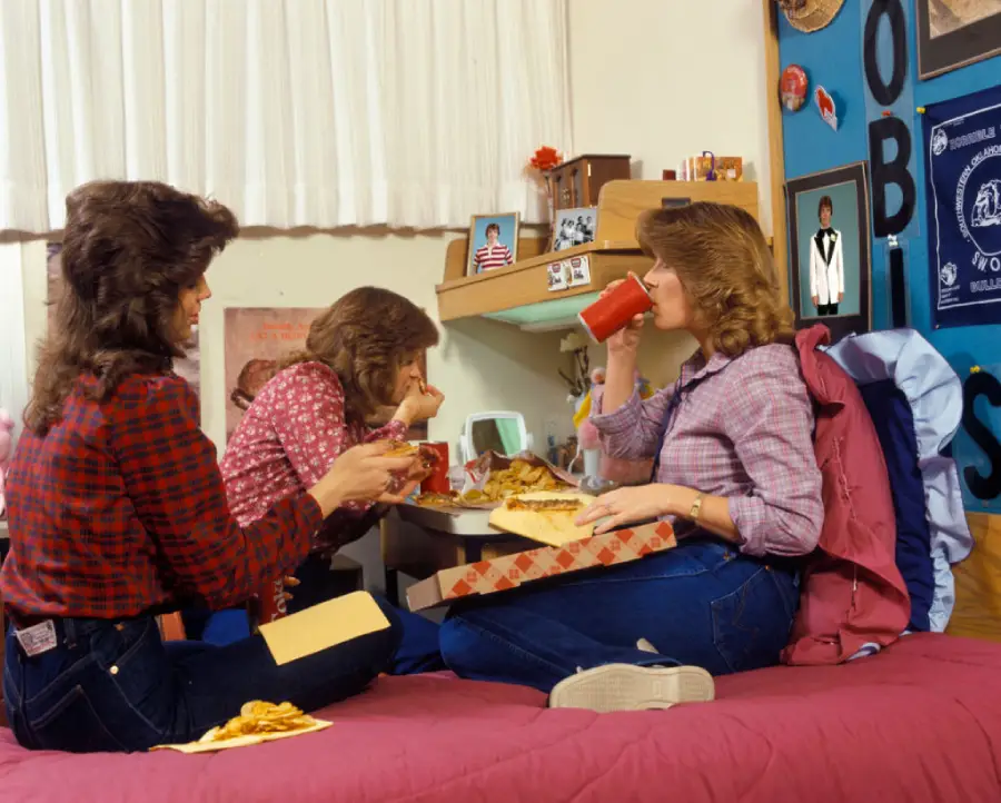 Three women with 1980s hairstyles and plaid shirts eat pizza and drink from mugs in a dorm room decorated with photos, posters, and school memorabilia. One sits on the bed, two sit at a small desk.