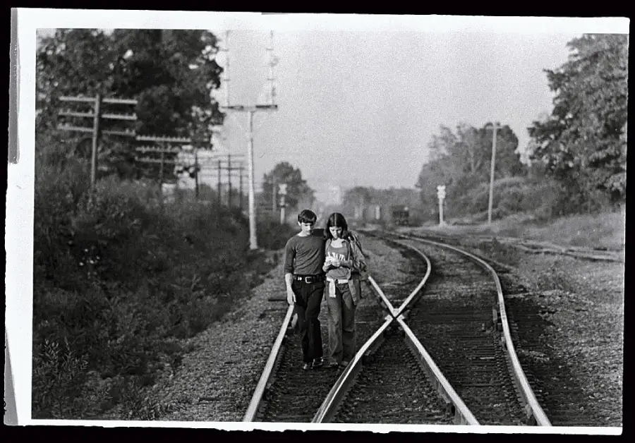 Two people walk closely together along curved railroad tracks, surrounded by trees and telephone poles, in a black and white outdoor scene.