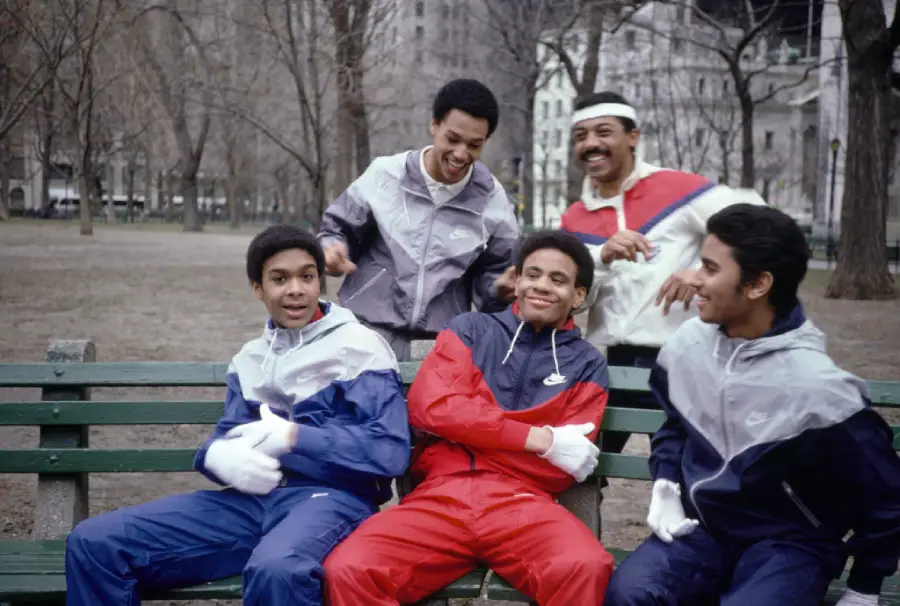 Four young men in colorful tracksuits sit and laugh on a park bench, while an older man in sportswear stands behind them, smiling. Leafless trees and city buildings are visible in the background.