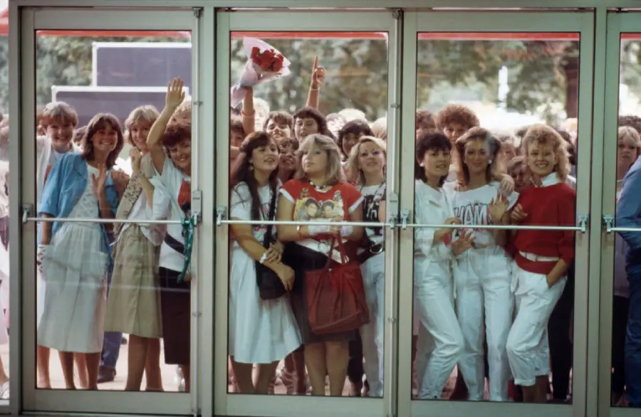 A group of excited young people, mostly women, stand behind glass doors, some waving and smiling, eagerly looking inside. They are dressed in casual 1980s clothes with bright colors and printed shirts.
