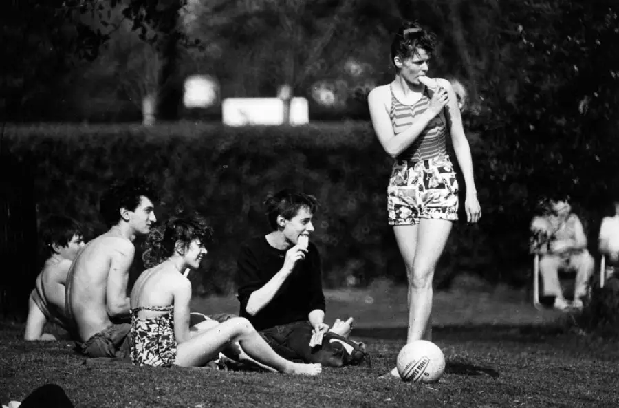 A group of young people relax on grass, some sitting and eating ice cream, while a woman in shorts stands nearby with a volleyball at her feet. The scene appears casual and summery, with trees and people in the background.