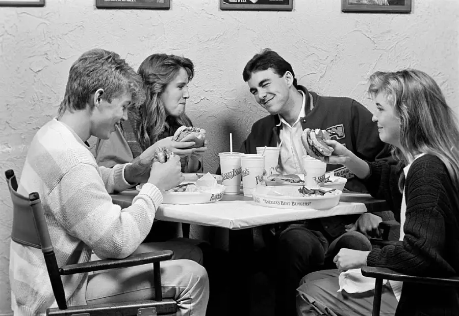 Four young adults sit around a table, smiling and eating burgers. There are drinks on the table, and the casual scene suggests they are enjoying a meal together at a restaurant. The image is in black and white.