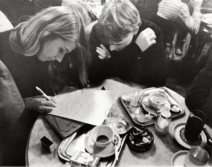 Two people sit at a table in a café. One writes on paper while the other watches. There are coffee cups, dishes, and trays on the table. Other patrons are visible in the background. The image is black and white.