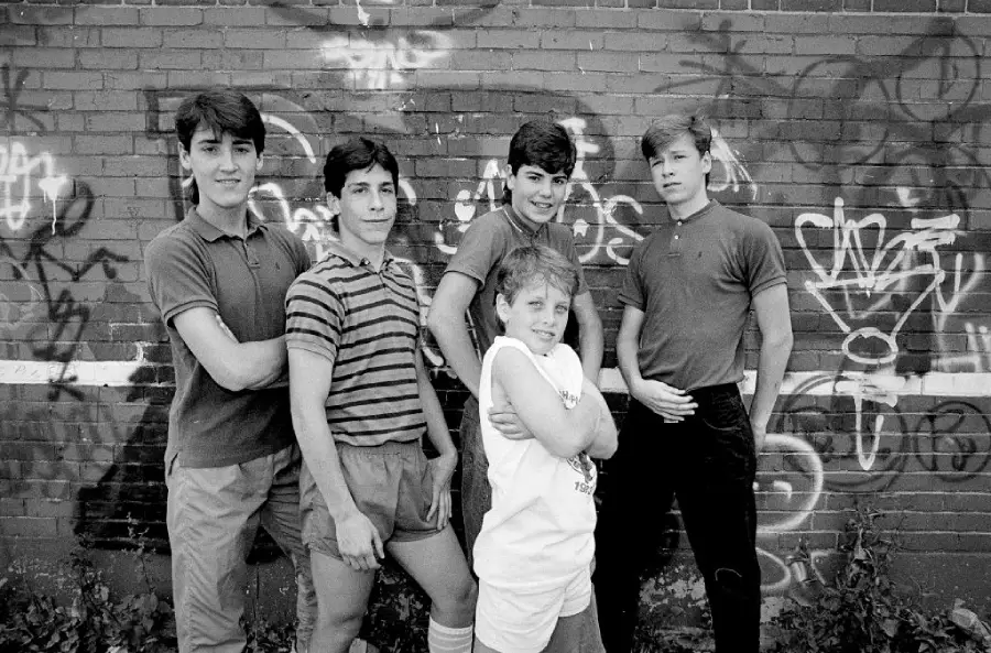 Five boys posing in front of a graffiti-covered brick wall, some with arms crossed and some with hands on their hips, all looking at the camera.