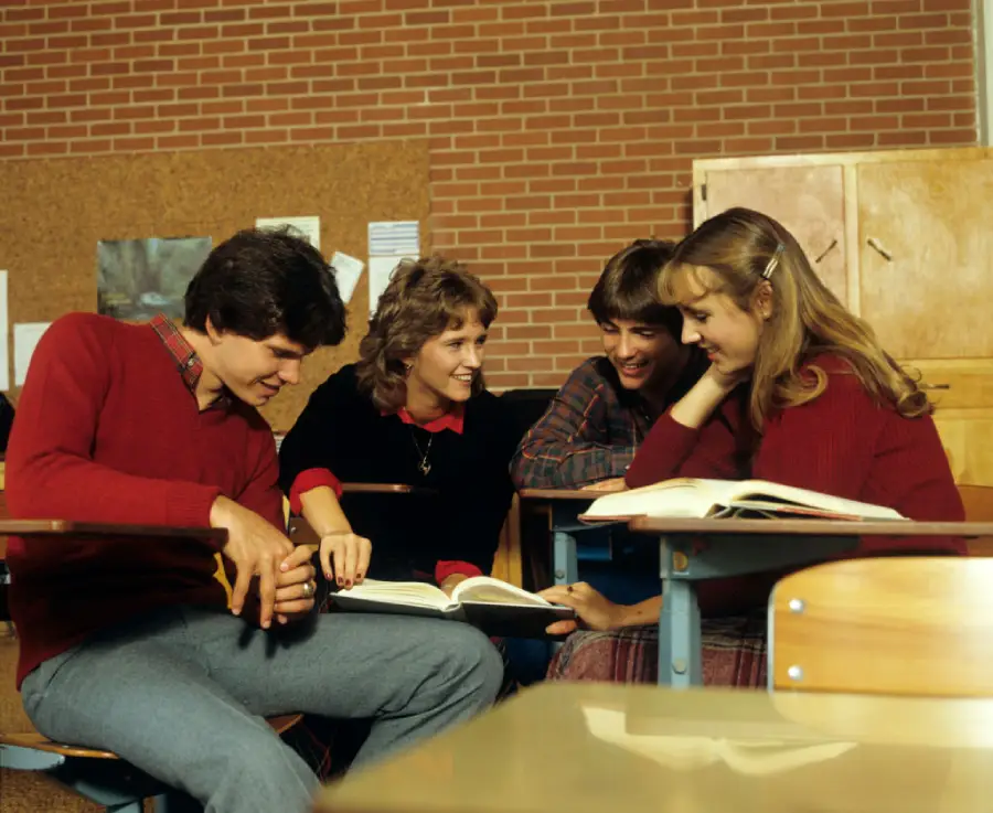 Four students sit together at desks in a classroom, smiling and talking while looking at open books. There is a brick wall and bulletin board in the background.