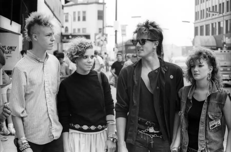 Four young people with punk hairstyles and edgy 1980s fashion stand together on a city street, looking in different directions. The photo is black and white, capturing an urban scene in daytime.