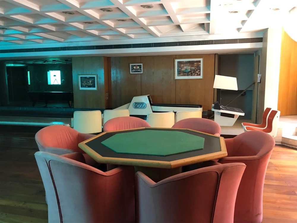 A room with a green octagonal card table surrounded by pink chairs, a white piano, air hockey table, wood panel walls, and framed pictures.
