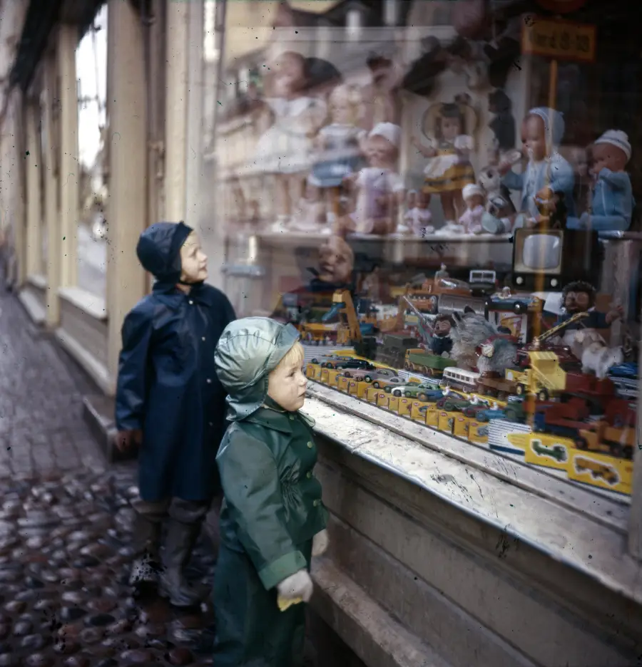 Two children in raincoats stand on a cobblestone street, gazing through a shop window filled with various toys, including dolls, stuffed animals, and colorful vehicles.