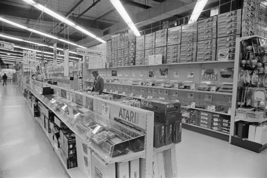 A black-and-white photo shows a large electronics store aisle filled with Atari video game consoles, accessories, and software, with one person browsing the home computer section. Bright lights line the ceiling above.