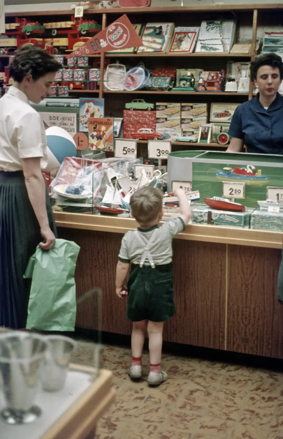 A young child points at toys in a glass display case while standing beside an adult, with a shopkeeper behind the counter in a toy store.