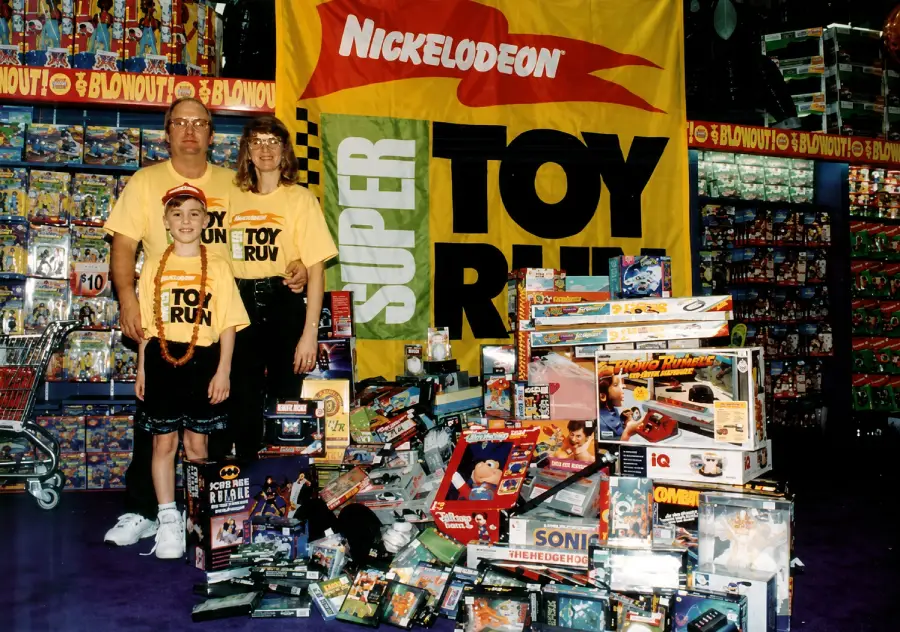 A family in yellow “Super Toy Run” shirts stands beside a large pile of toys in front of a Nickelodeon Super Toy Run banner, with toy store shelves in the background.