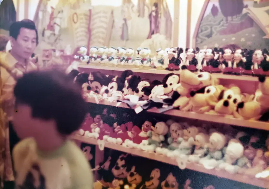 A man stands near shelves filled with various plush toys, while a child in a green shirt walks by in the foreground. The background includes colorful wall art.