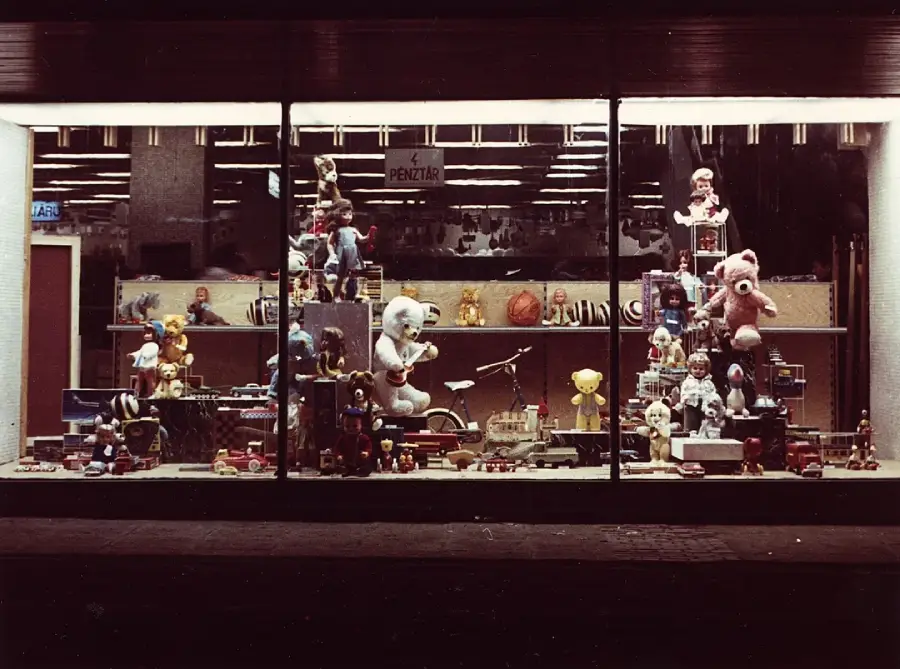 A toy store window display at night features stuffed animals, dolls, toy vehicles, and various toys arranged on shelves with a PENZTA sign above. The scene is warmly lit from inside.