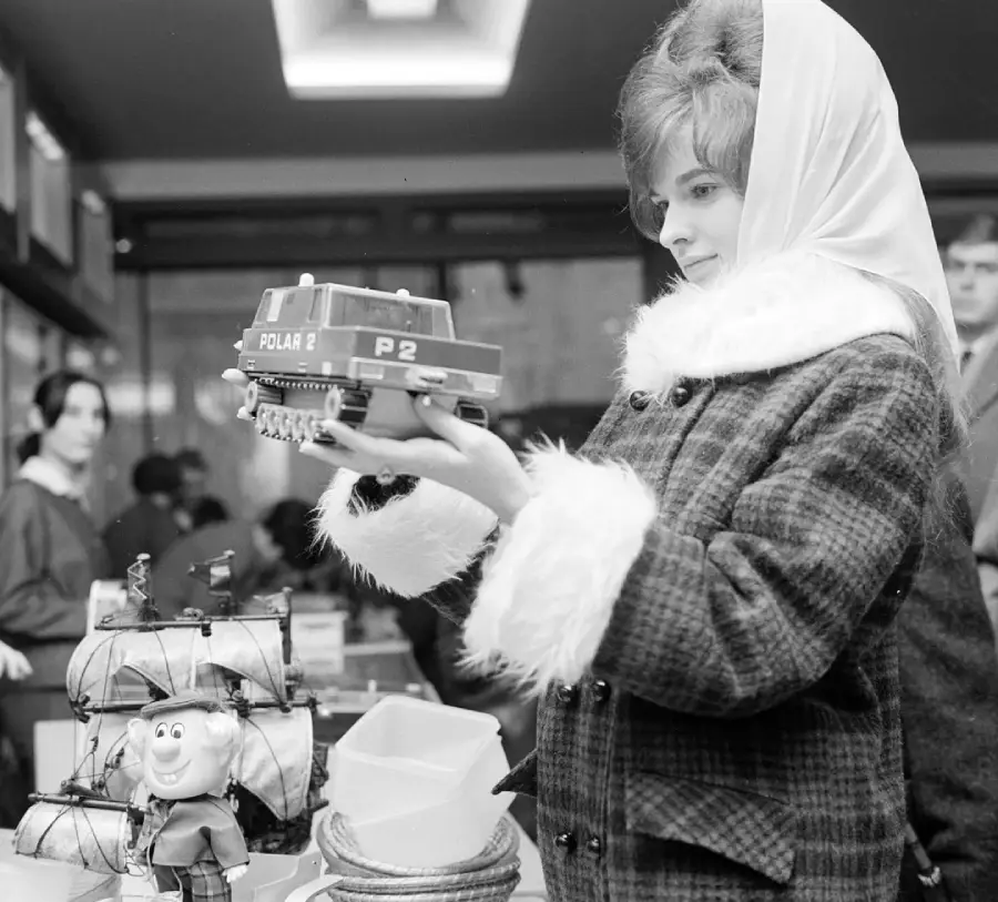 A woman in a fur-trimmed coat and headscarf holds a toy vehicle in a store. Toys, including a ship and a doll, are on the counter. Other shoppers are visible in the background.
