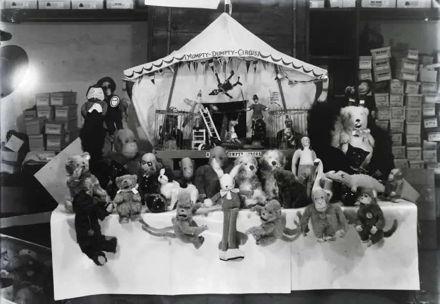 A black-and-white photo of various vintage dolls, teddy bears, and toys arranged on a table in front of a toy circus tent labeled “Humpty-Dumpty Circus,” with shelves of boxes in the background.