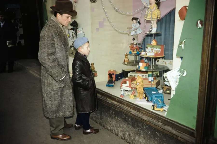 A man and a young boy in winter coats and hats look at a toy store window display filled with dolls, toy cars, and holiday decorations.