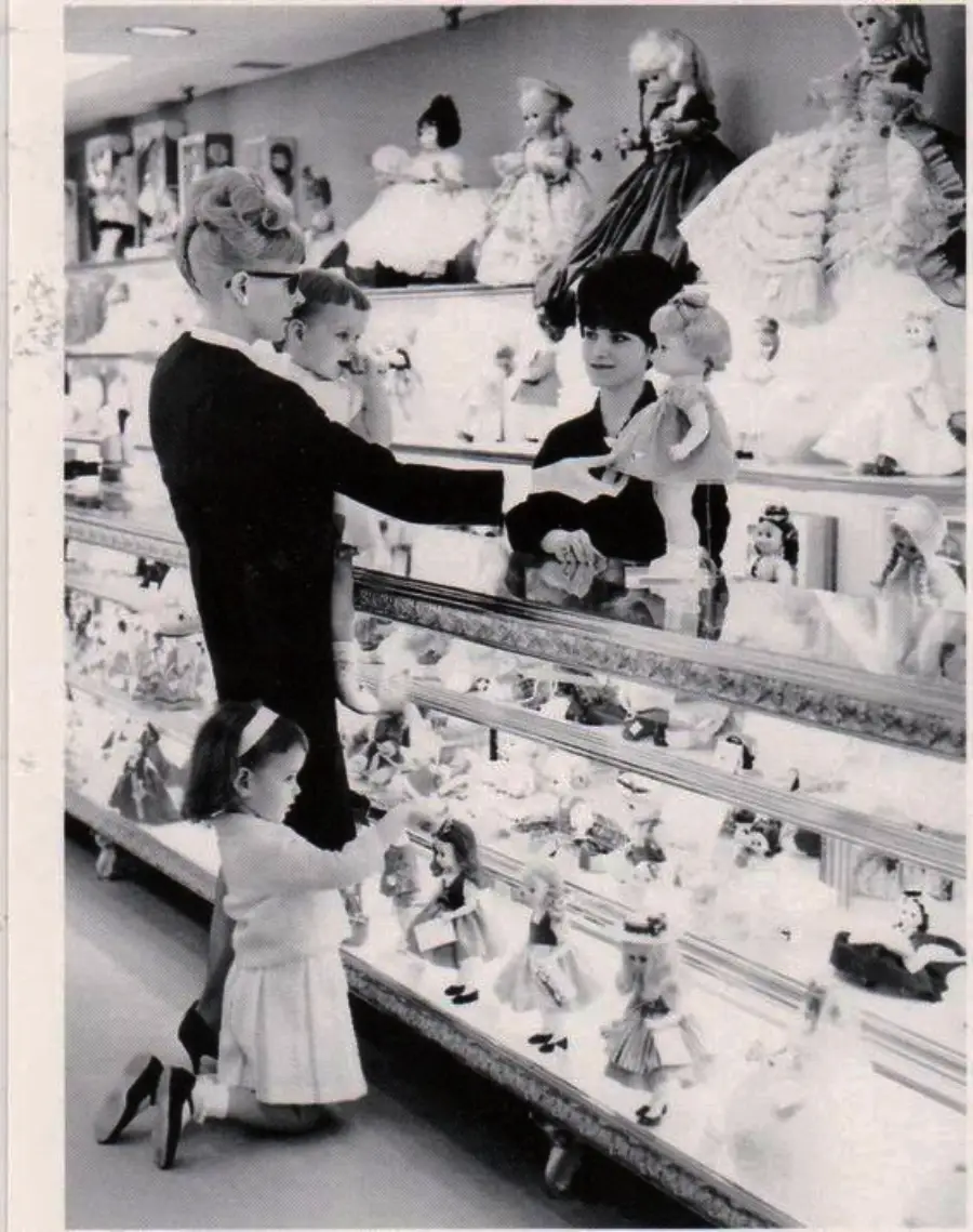 A woman holding a baby and a small girl stand at a glass counter filled with dolls, while a shop worker shows them a doll. More dolls are displayed on top of the counter and shelves behind.