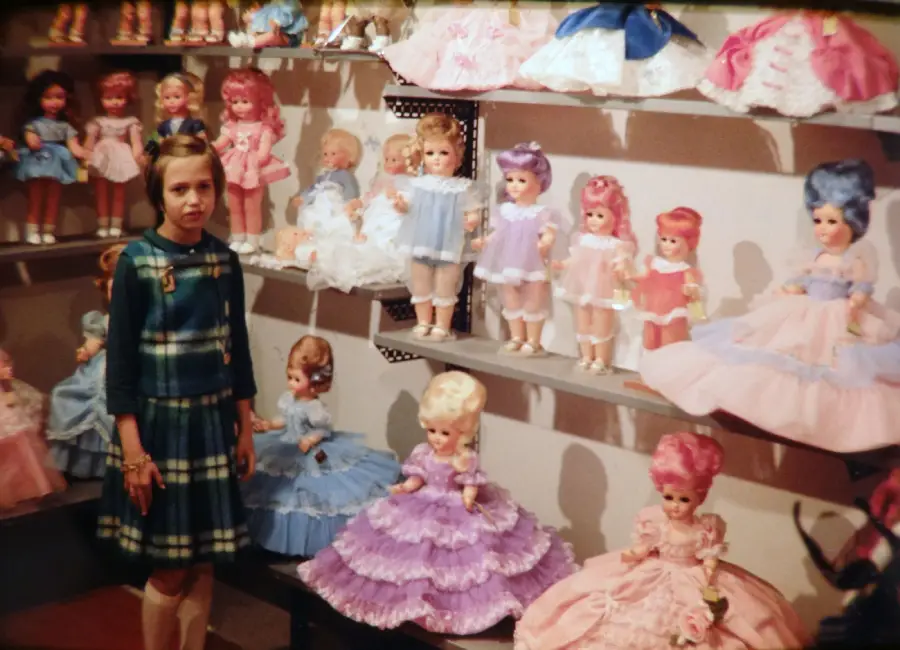 A young girl in a green plaid dress stands in front of shelves filled with large, elaborately dressed dolls wearing colorful, ruffled gowns.